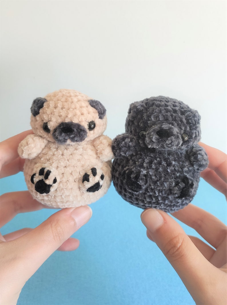 May include: Two crocheted plush toys, one beige and one gray, shaped like pugs. Both have black eyes and noses.