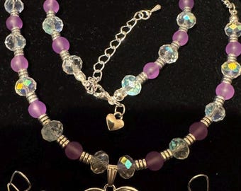 Puede incluir: Un conjunto de collar y pendientes de cristal morado y transparente con un colgante de corazón doble. El collar tiene una cadena de plata y un dije de corazón.