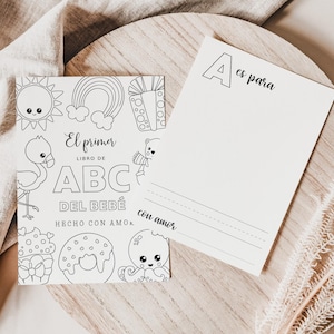 Könnte beinhalten: Ein Malbuch und eine leere Karte werden auf einer Holzoberfläche gezeigt. Das Malbuch zeigt Strichzeichnungen einer Sonne, eines Regenbogens und anderer Bilder mit dem Text "El primer libro de ABC del bebé hecho con amor." Die leere Karte hat den Buchstaben "A" und die Worte "es para" und "con amor."