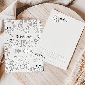 May include: A black and white coloring book titled "Baby's first ABC Book" with illustrations of a sun, rainbow, gift, flamingo, cupcake, donut, and octopus. A matching card reads "A is for" with lines for writing and the words "with love".