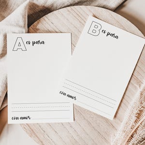 Könnte beinhalten: Zwei weiße Karten mit schwarzem Text, eine mit dem Buchstaben "A" und den Worten "es para" und "con amor", die andere mit "B" und "es para" und "con amor". Die Karten liegen auf einem Holztablett mit beigem Stoff und einer getrockneten Pflanze.
