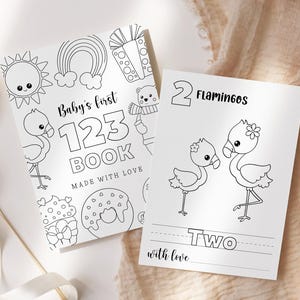 May include: Two black and white illustrated pages from a children's book. One page reads "Baby's first 123 Book" with illustrations of a sun, rainbow, flamingo, and other objects. The other page shows two flamingos and the word "Two".