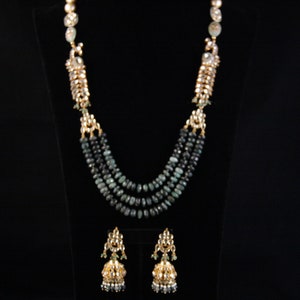 May include: A three-strand necklace and matching earrings set. The necklace features dark green beads and gold-colored accents. The earrings have a bell shape with gold filigree and green accents. The jewelry is displayed against a black background.