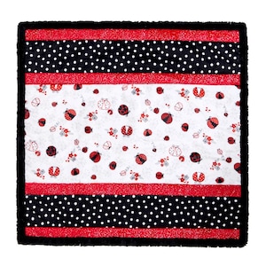May include: Square quilt featuring a white center with red ladybug and floral patterns. The quilt has red and black polka dot borders, red stripes, and a black fringed edge. A cozy, decorative item.