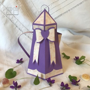 Può includere: Una teiera di carta viola con un designo a motivi bianchi e rosa. La teiera ha un fiocco con un cuore al centro. La teiera è appoggiata su una superficie bianca con fiori viola.