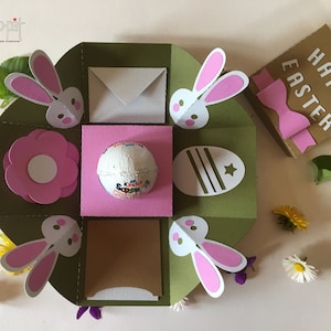 May include: A green and pink explosion box with a bunny rabbit theme. The box features a pink flower, a chocolate egg, and a white envelope. The box is decorated with a pink bow and the words "Happy Easter".