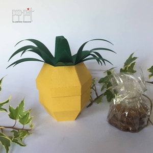 May include: A yellow pineapple-shaped paper box with a green paper top and a green paper handle. The box is sitting on a white surface. The text "pop-pat by patrizia moscone" is printed on the box.