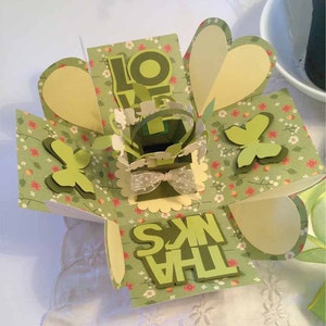 以下が含まれることがあります： 前面に「ありがとう」と「愛」の文字が書かれた緑と黄色のポップアップボックスカード。カードは蝶と花で飾られています。