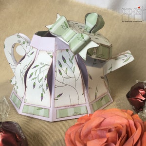 May include: A white and green paper teapot with a purple lid and a green bow. The teapot is decorated with a floral pattern and has a scalloped edge. The teapot is sitting on a brown surface.
