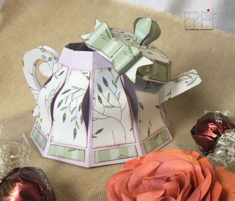 May include: A white and green paper teapot with a purple lid and a green bow. The teapot is decorated with a floral pattern and has a scalloped edge. The teapot is sitting on a brown surface.