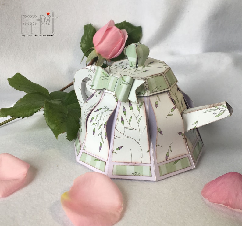 May include: A white paper teapot with a green and purple floral pattern and a green bow. The teapot is sitting on a white surface with pink rose petals.