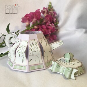 May include: A green and white paper teapot with a floral pattern and a matching lid. The teapot is open and the lid is off. The teapot is sitting on a white surface.