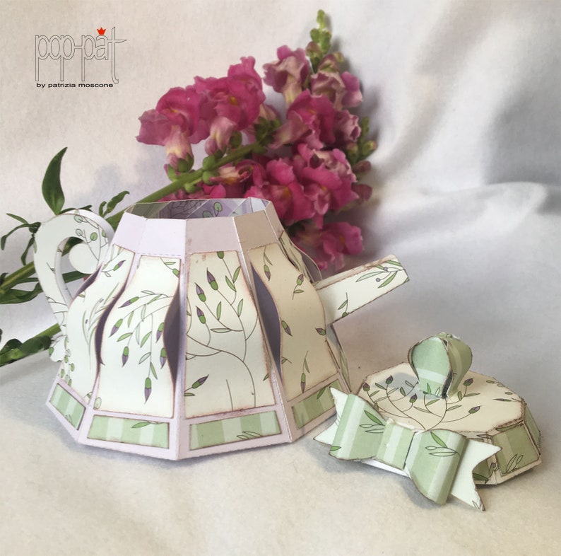 May include: A green and white paper teapot with a floral pattern and a matching lid. The teapot is open and the lid is off. The teapot is sitting on a white surface.