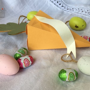Può includere: Una scatola di carta arancione a forma di carota con un nastro bianco e una coda di coniglio di carta verde. La scatola è piena di uova di Pasqua di cioccolato e si trova su una superficie di tessuto bianco.