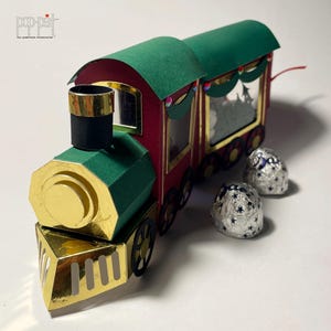 May include: A handcrafted paper train with a red body, green roof, and gold accents. The train features a decorative design and is accompanied by two foil-wrapped candies. The image includes the text "pop-pat" by patricia moscone.