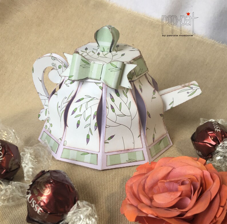 May include: A white paper teapot with a green and white striped bow, green leaves, and purple accents. The teapot is a pop-up box, designed by Patricia Moscone.