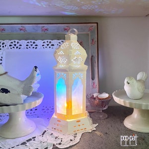 Op de afbeelding: Een witte papieren lantaarn met ingewikkelde uitsnijdingen en een blauw en geel licht dat erdoorheen schijnt. De lantaarn staat op een witte kantonderlegger en een witte tafel.