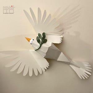 May include: A white paper dove with a green olive branch in its beak, flying with its wings spread wide. The dove is a symbol of peace and is made of paper.