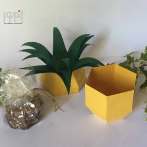 May include: A yellow hexagonal box with a green pineapple top. The box is open and empty. The pineapple top is made of paper and has a green color.