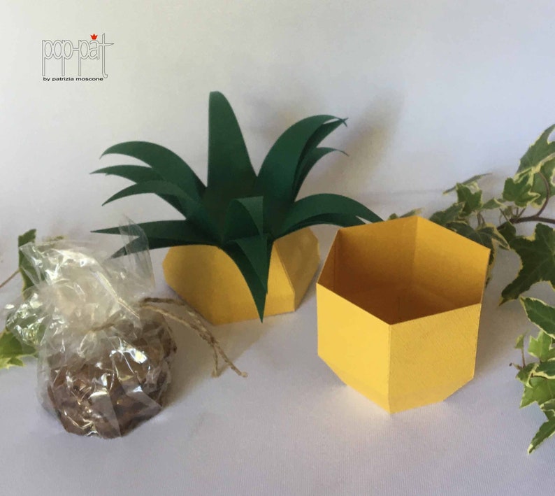 May include: A yellow hexagonal box with a green pineapple top. The box is open and empty. The pineapple top is made of paper and has a green color.