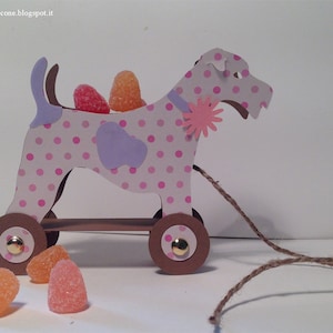 Può includere: Un cane giocattolo rosa e bianco a pois con orecchie viola e un fiore rosa sul colletto. Il cane ha ruote marroni e tira una corda. Ci sono caramelle arancioni e rosa sul pavimento.