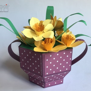 May include: A purple and white polka dot papercraft teacup with a handle, filled with yellow paper daffodils and green paper leaves. The teacup is a 3D paper craft.