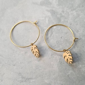 May include: A pair of gold hoop earrings with a small, cutout monstera leaf charm hanging from each hoop.