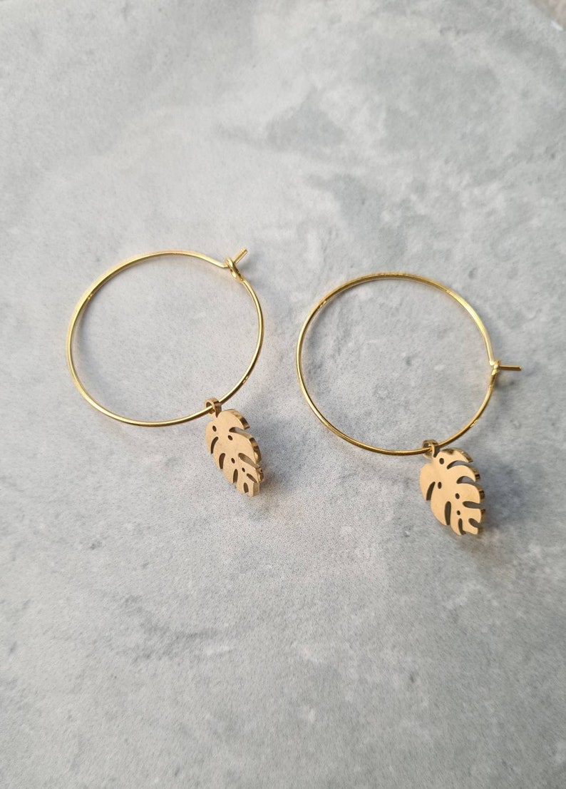 May include: A pair of gold hoop earrings with a small, cutout monstera leaf charm hanging from each hoop.