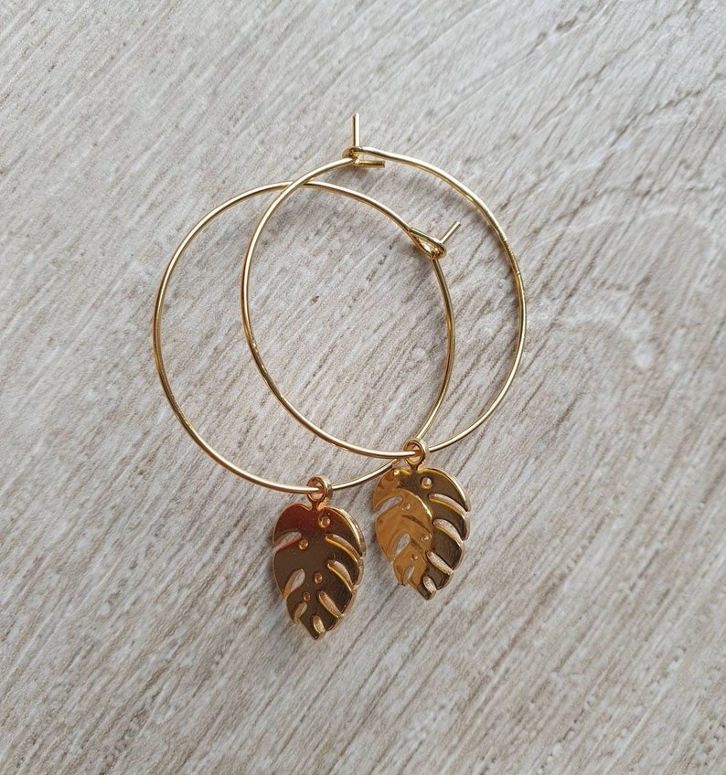 May include: Gold hoop earrings with a gold monstera leaf charm hanging from each hoop.