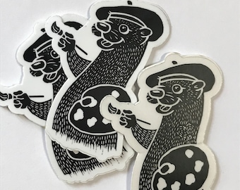 May include: A stack of black and white stickers featuring a cartoon otter wearing a beret and holding a paintbrush and artist's palette. The otter has a smiling face and is depicted in a detailed, linework style.