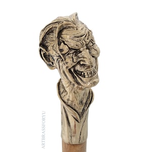 May include: A brass cane handle sculpted into the detailed head of a grinning character with slicked-back hair and prominent facial features. The metal has a polished, aged finish with dark recessed areas. The text ARTBRASSFORYU is visible.