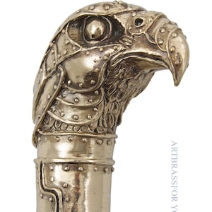 May include: A detailed, golden-colored walking cane handle shaped like a stylized eagle's head. The eagle's head is adorned with rivet-like details, giving it a textured, metallic appearance. The handle is a decorative accessory.