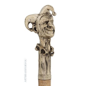 May include: A silver-toned walking cane with a detailed jester head handle. The jester has a smiling expression and a pointed hat. The cane's shaft is a light brown color, and the handle is intricately sculpted.
