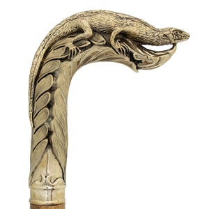 May include: A decorative walking cane with a detailed lizard figure atop the curved handle. The handle is a light gold color with leaf-like carvings. The lower portion of the cane is a light brown color.
