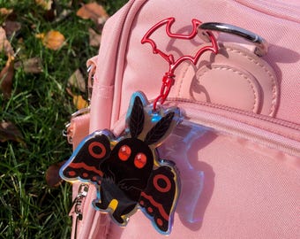 May include: A black moth charm with red eyes and wing details, attached to a red bat-shaped keychain. The charm is hanging from a pink backpack, set against a backdrop of green grass and fallen leaves.