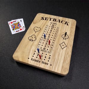 May include: A wooden SETBACK board game with engraved suit symbols and scoring tracks. The board has blue and red pegs, with the text "SETBACK" and "GAMES WON". Two playing cards are visible.