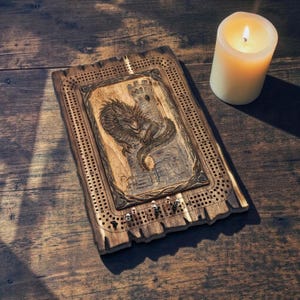 May include: A rectangular wooden cribbage board featuring a carved dragon and castle design in the center. The board has rows of drilled holes for scoring, with small metal pegs inserted near the start and win positions on the game track.