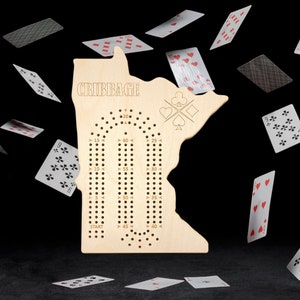 May include: A wooden cribbage board in the shape of a state, with the word "CRIBBAGE" at the top. The board has holes for pegs and is surrounded by playing cards. The board is made of light-colored wood.