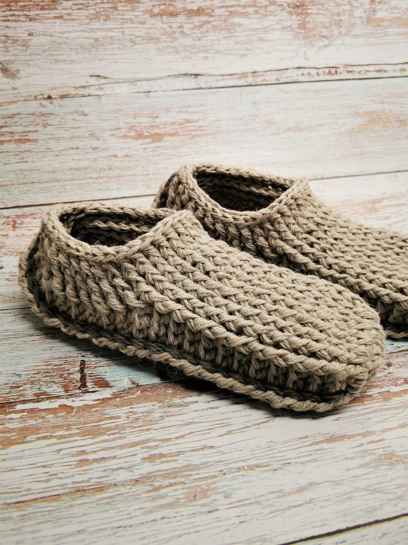 May include: A pair of beige crocheted slippers on a whitewashed wooden surface.