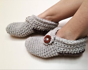 Può includere: Un paio di pantofole in crochet grigio con dettagli in bottone marrone. Le pantofole sono indossate da una persona seduta su una superficie bianca.