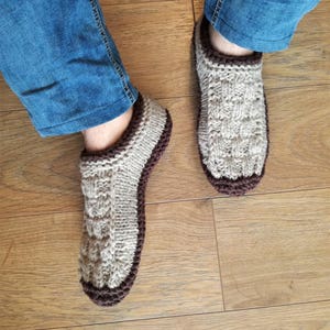 May include: A pair of knitted beige slippers with brown trim. The slippers are worn on a wooden floor.