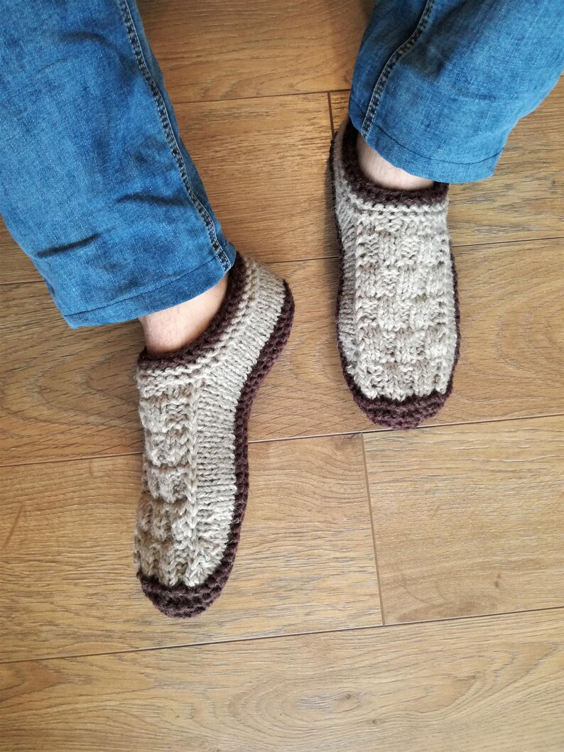 May include: A pair of knitted beige slippers with brown trim. The slippers are worn on a wooden floor.
