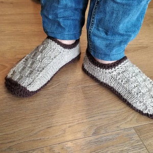 May include: A pair of knitted slippers with a brown trim. The slippers are made of a light gray yarn and have a cable knit pattern.