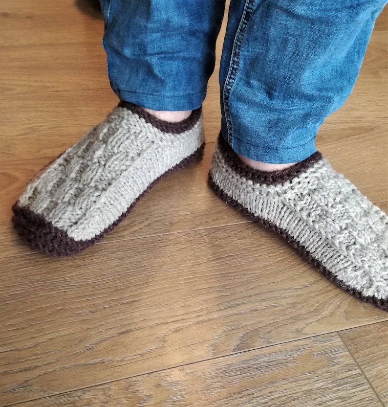 May include: A pair of knitted slippers with a brown trim. The slippers are made of a light gray yarn and have a cable knit pattern.