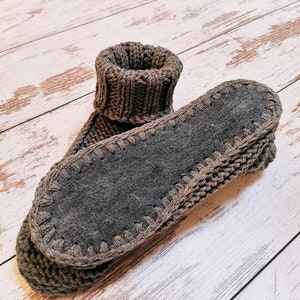 May include: A pair of gray knitted slipper socks with a brown suede sole. The socks are folded over and the soles are facing the camera.