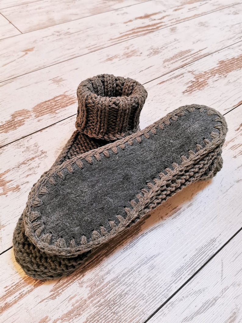 May include: A pair of gray knitted slipper socks with a brown suede sole. The socks are folded over and the soles are facing the camera.