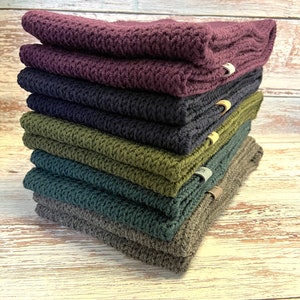 Puede incluir: Una pila de seis bufandas de punto en varios colores, incluyendo morado, azul oscuro, verde y gris. Las bufandas están dobladas y apiladas sobre una superficie de madera.