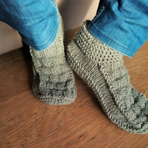 May include: A pair of gray knitted slipper socks with a cable knit pattern. The socks are worn on a person's feet with blue jeans.