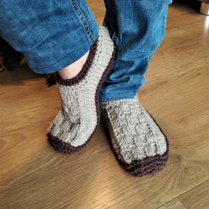 May include: A pair of knitted slippers with a brown trim. The slippers are made of a light gray yarn and have a cable knit pattern.
