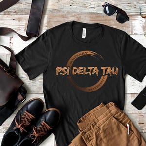May include: Black t-shirt with the text "MILITARY PSI DELTA TAU FRATERNITY" in brown lettering and a circular design. The shirt is displayed with brown pants, black shoes, a leather bag, and sunglasses.